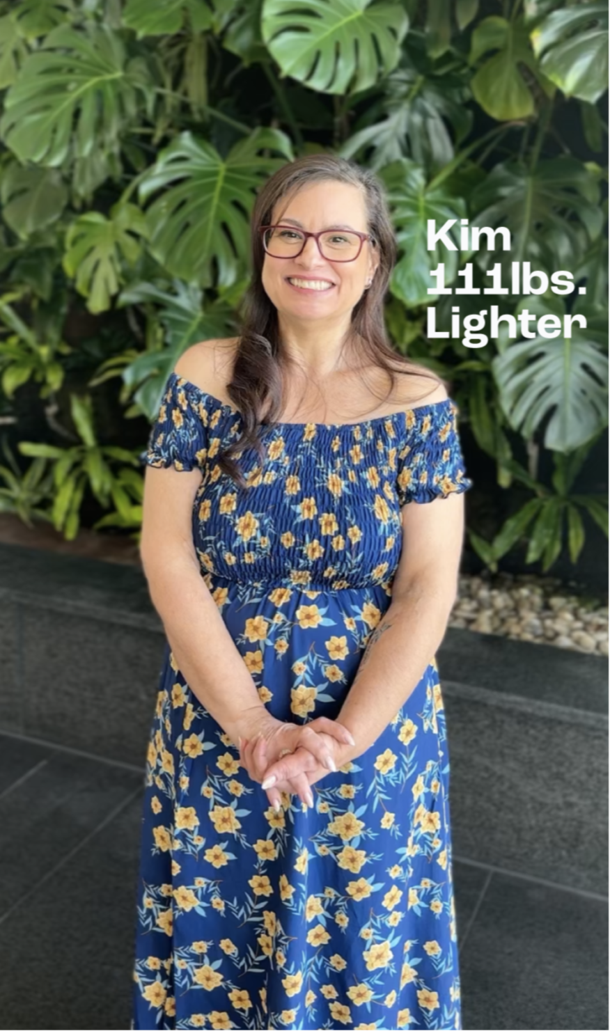 Woman in a blue floral dress standing in front of green foliage with text 'Kim 111lbs. Lighter'.