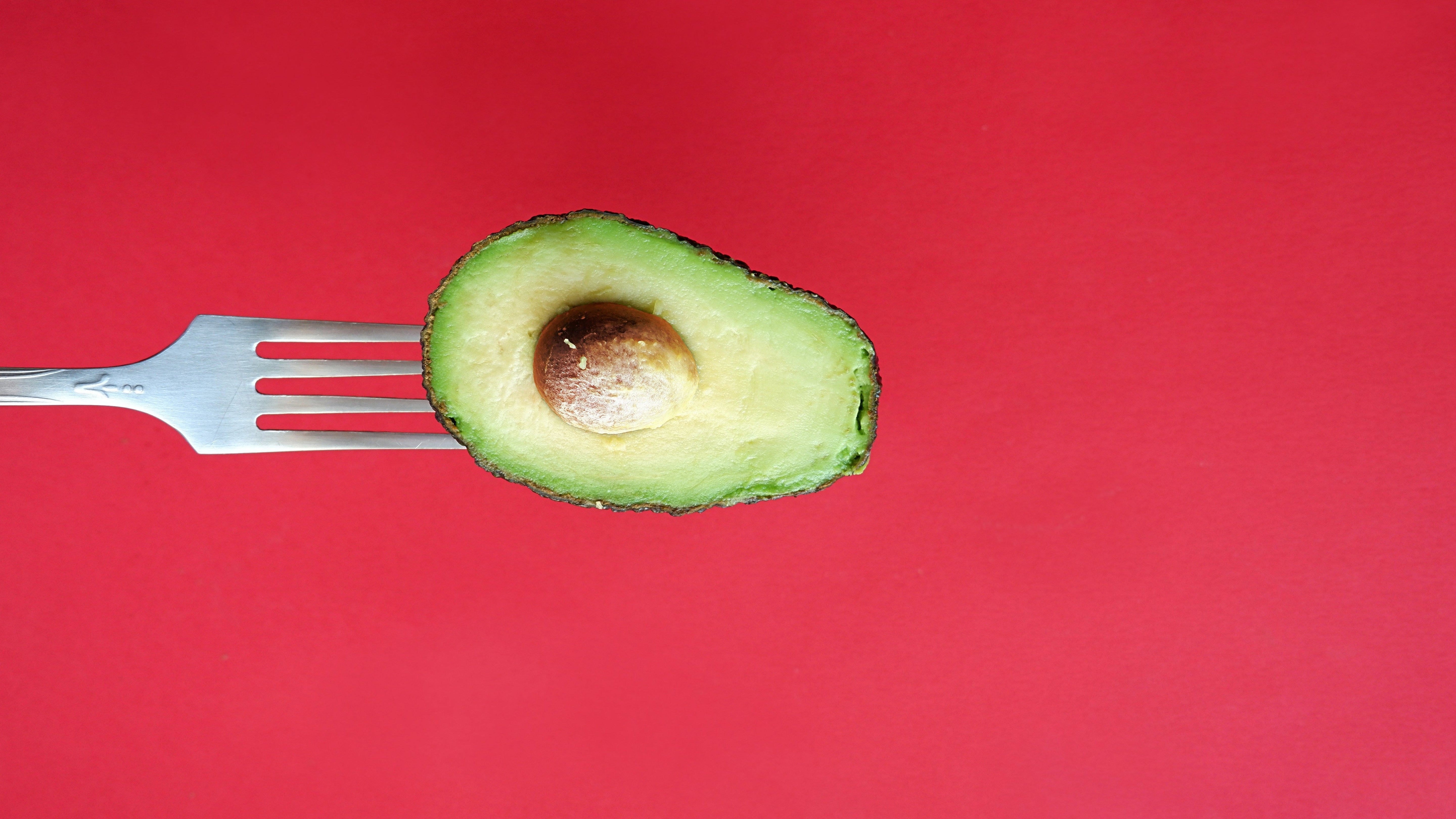 Half of an avocado on a fork against a red background