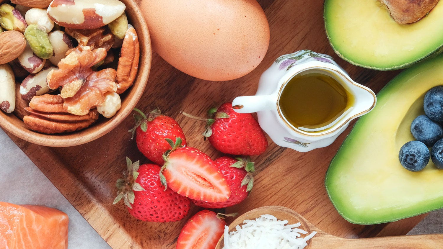 Assorted healthy foods including nuts, eggs, avocados, berries, and salmon on a wooden surface.