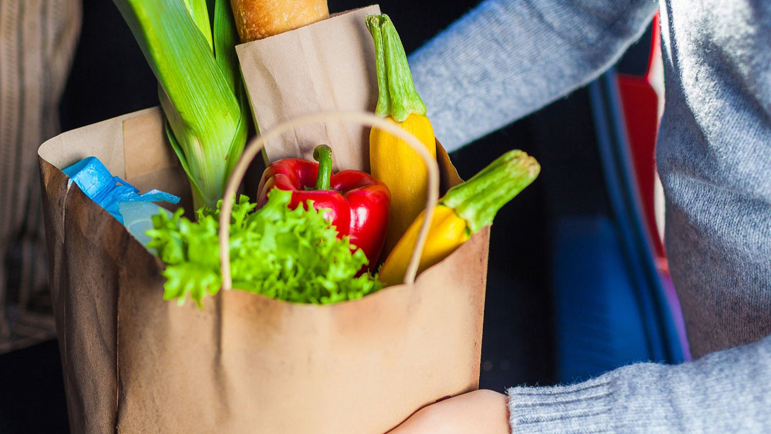 Person holding a brown paper bag filled with groceries including vegetables and a loaf of bread.