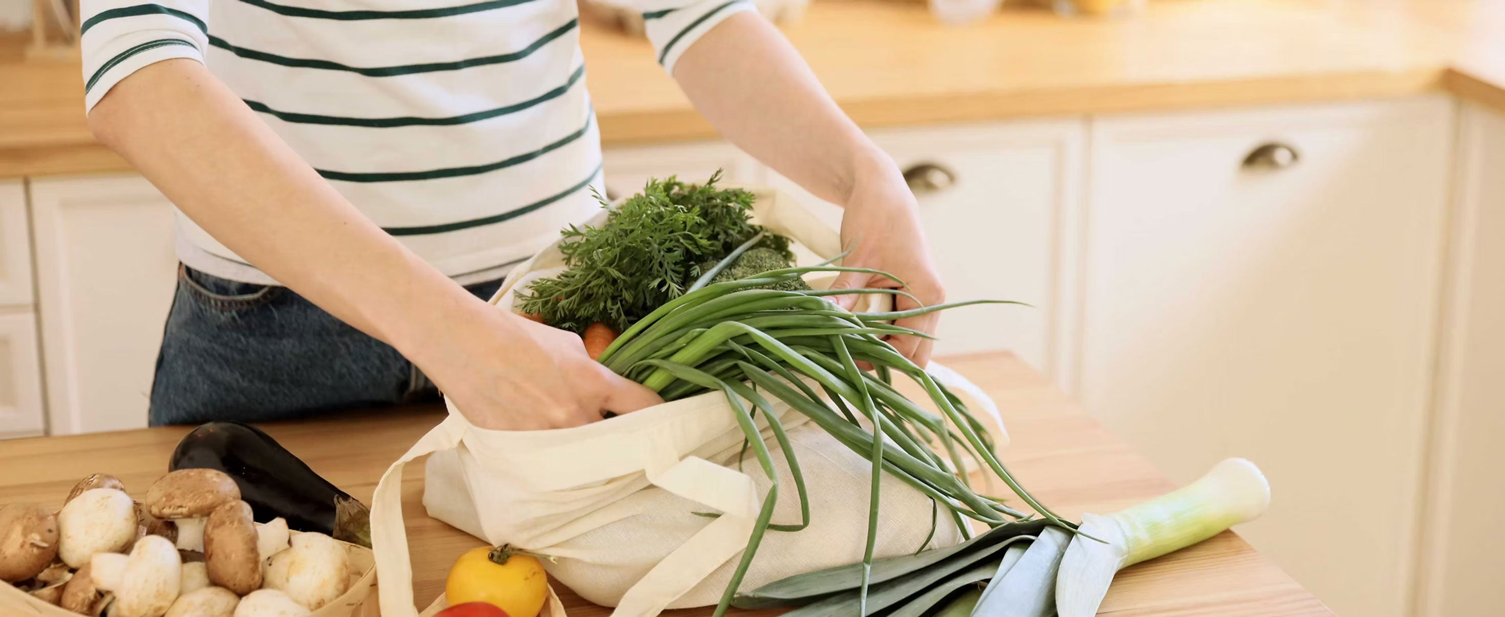 A woman removing leafy greens and vegetables from a canvas tote onto a kitchen counter.