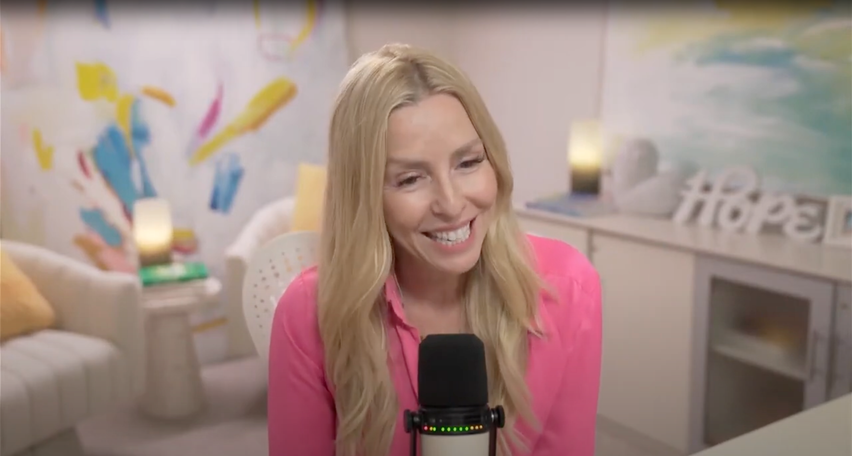 Gina Livy wearing a pink blouse, sitting in front of a microphone at her desk addresses the camera.