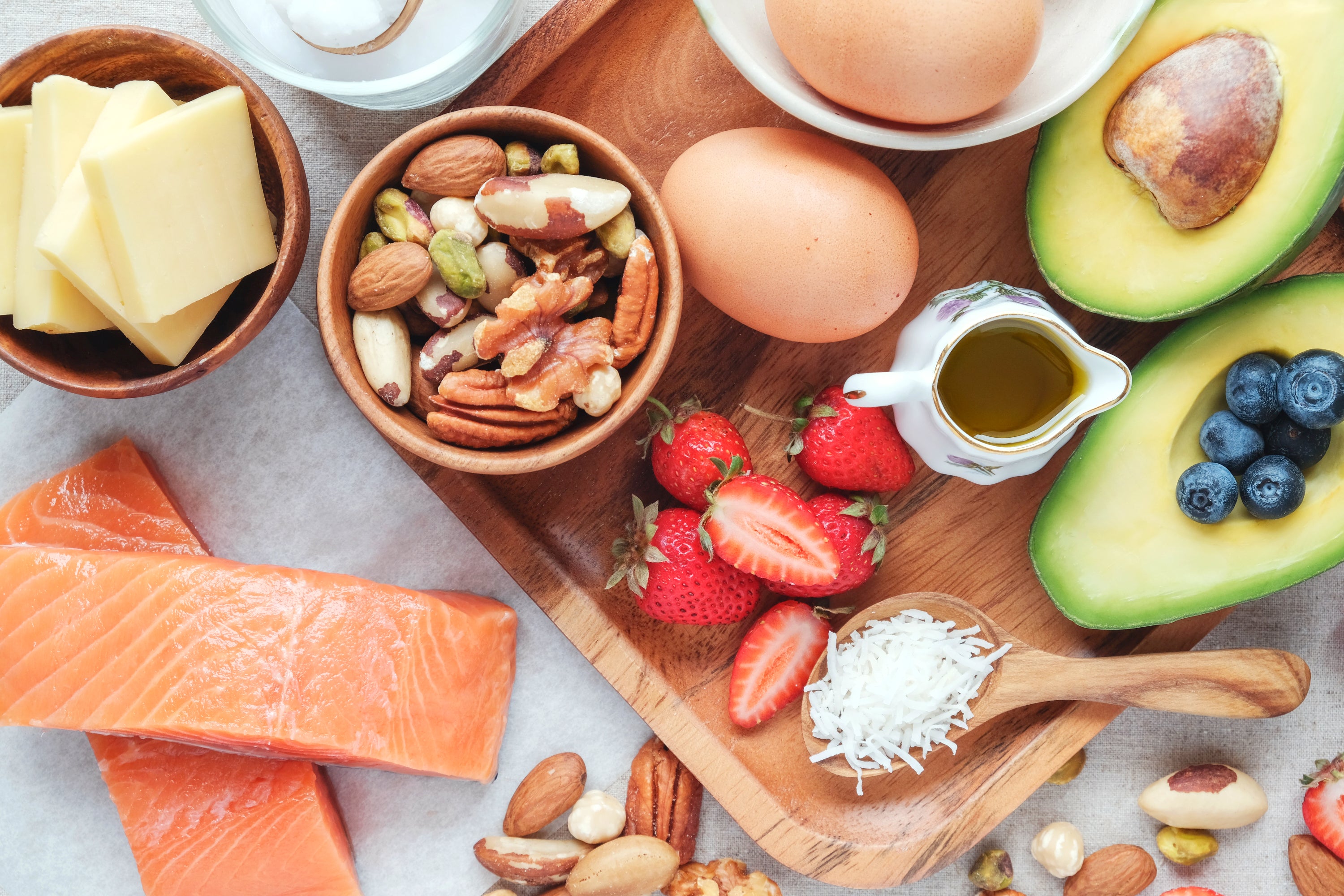 This image presents a flat lay composition showcasing a variety of healthy, whole foods, including avocados, berries, nuts, salmon, eggs, cheese and bowls of mixed nuts on a wooden cutting board—evoking fresh, mindful eating.