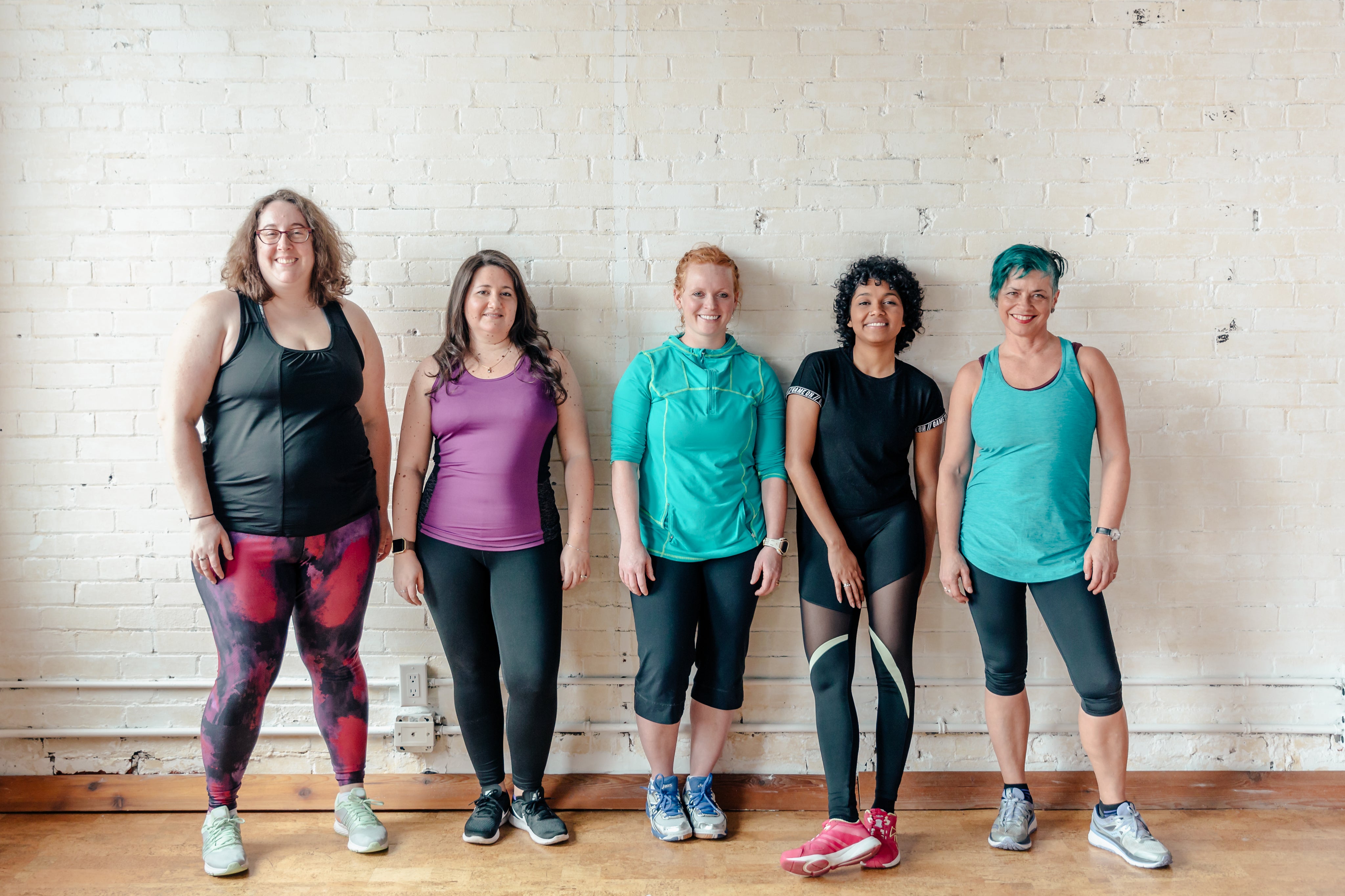 5 women dressed in workout attire standing against a white brick wall smiling