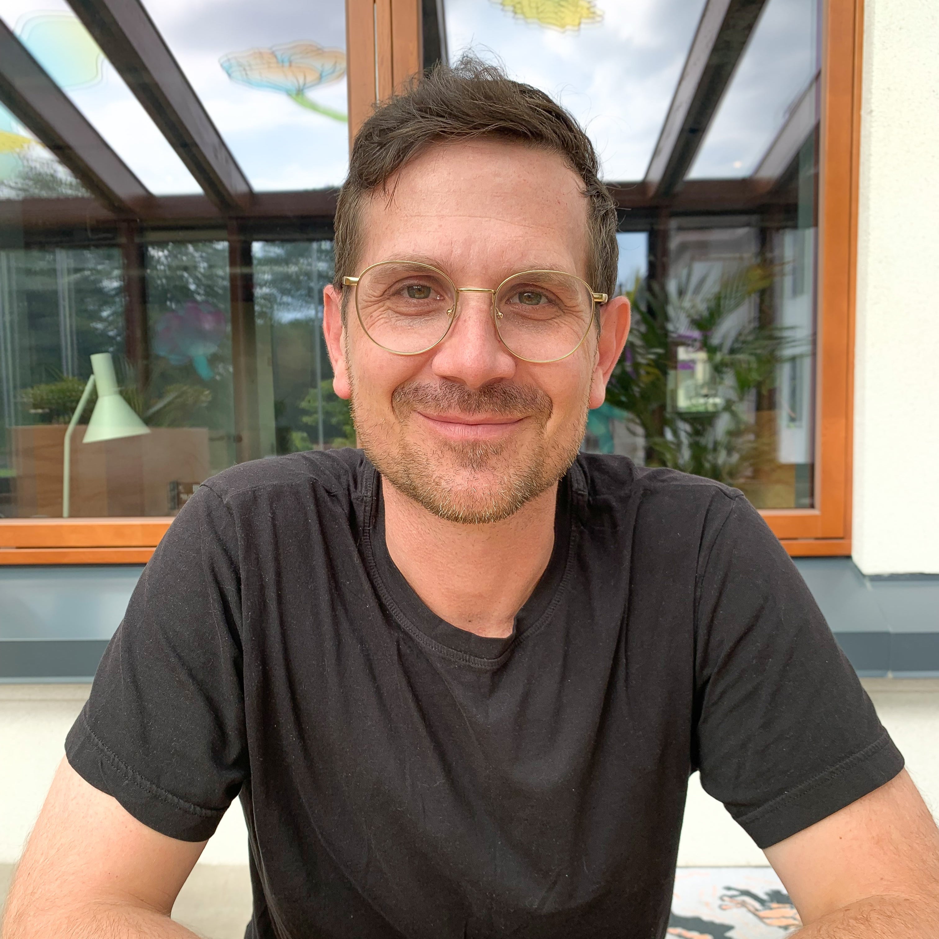 Jody Downdall wearing glasses and a black shirt sitting in front of a window with plants
